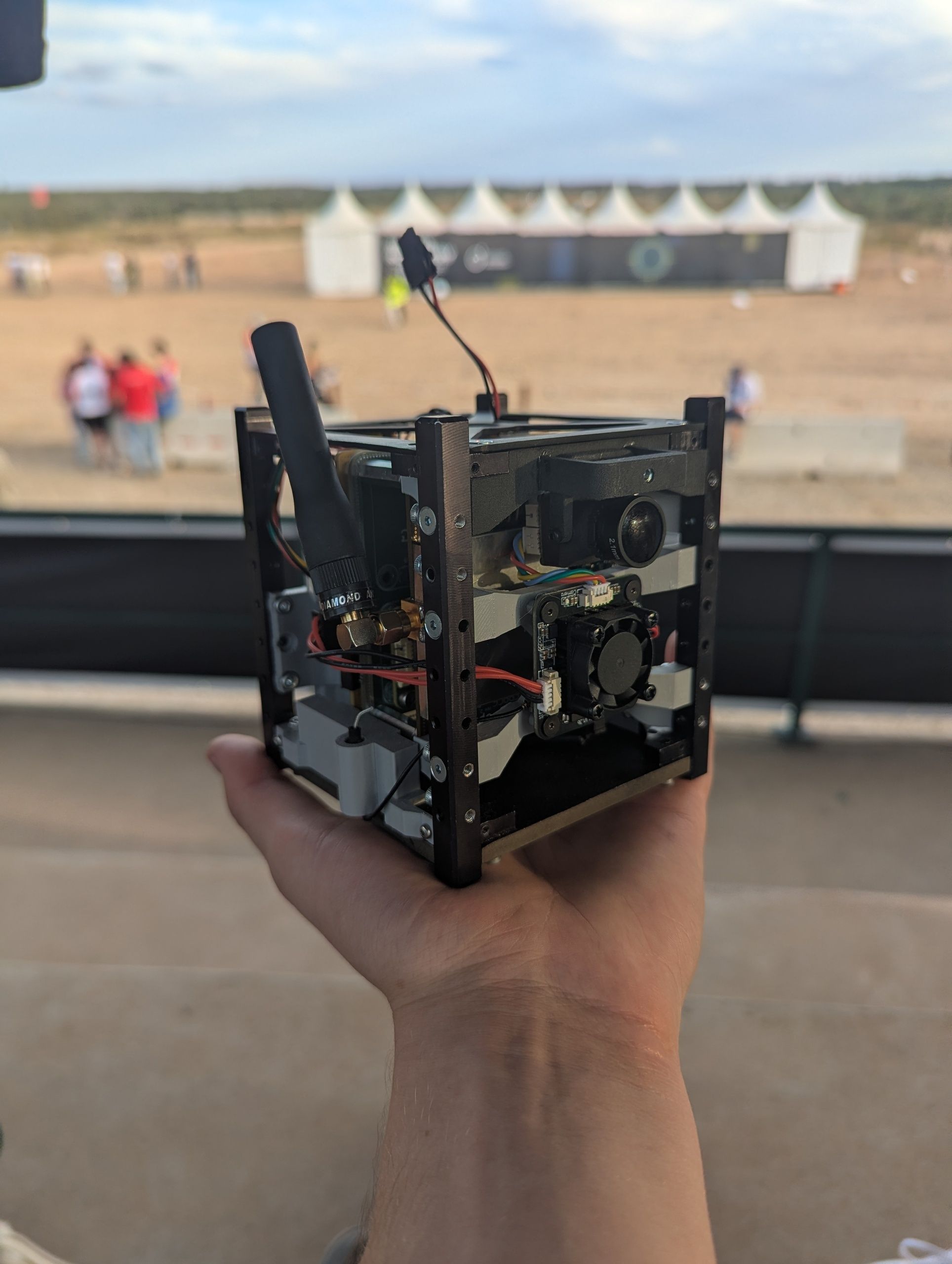 Hand holding the assembled South Cube payload at the EUROC launch site, with competition tents in the background